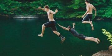 Three shirtless men jump into a green lake from a rocky ledge, midair in a sunny outdoor scene.