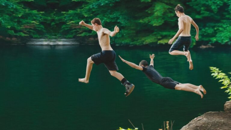 Three shirtless men jump into a green lake from a rocky ledge, midair in a sunny outdoor scene.
