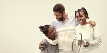 Father with his two daughters, smiling and embracing; the teen holds a tennis racket.